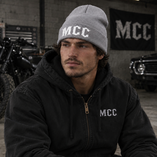 A man wearing a gray knit beanie with white 'MCC' lettering and a black zip-up hooded jacket with a matching 'MCC' logo on the left chest, posed in a garage setting with a motorcycle and car in the background.
