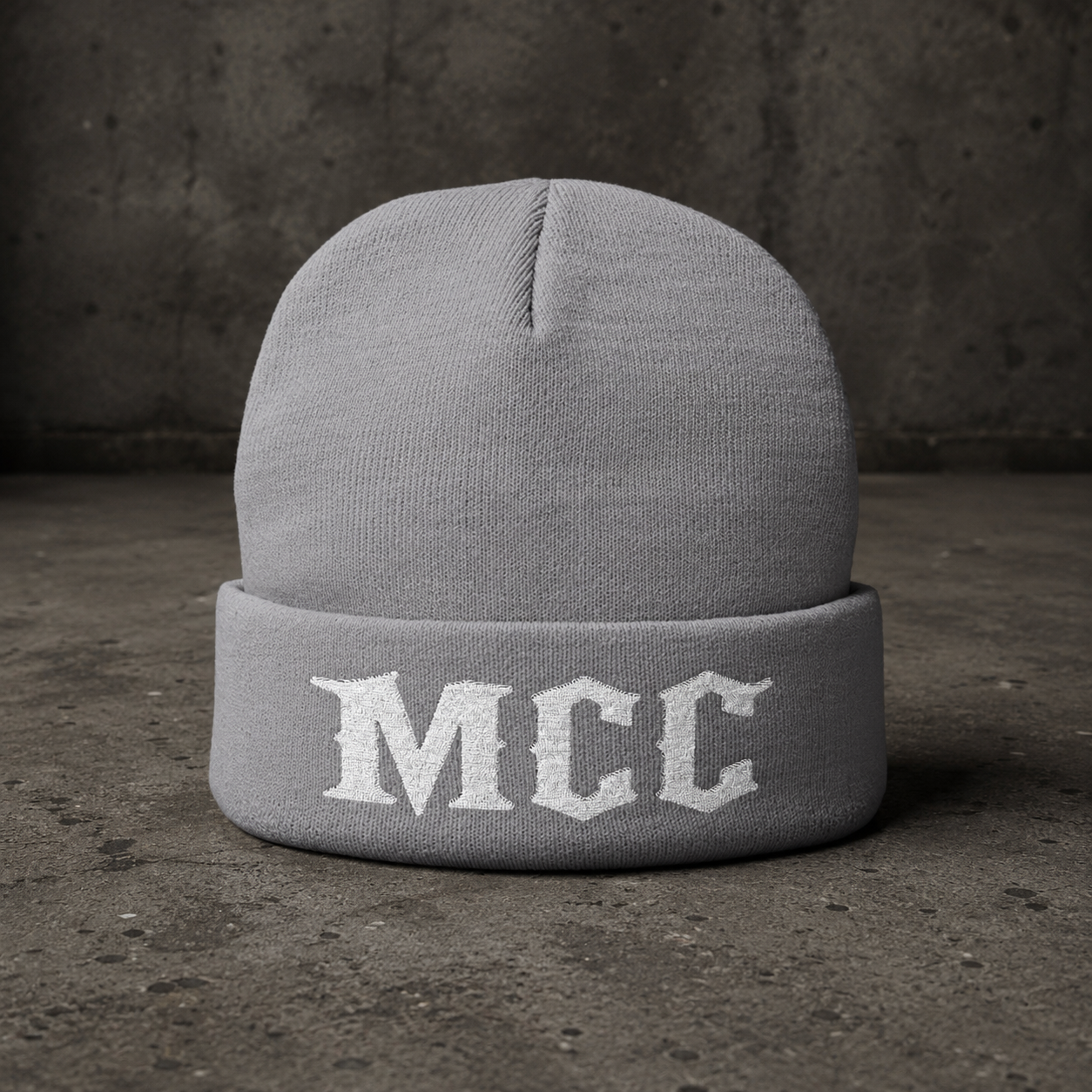 A light gray knit beanie hat with a folded cuff featuring the letters 'MCC' in white Gothic-style embroidery, set against a dark textured concrete background.