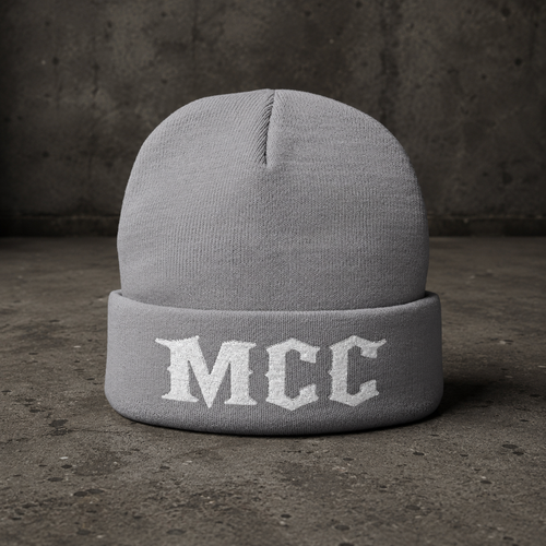 A light gray knit beanie hat with a folded cuff featuring the letters 'MCC' in white Gothic-style embroidery, set against a dark textured concrete background.