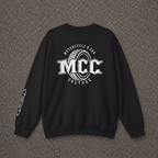 A black crewneck sweatshirt laid flat on a textured gray asphalt surface, featuring a large white graphic on the back with the text 'MCC Motorcycle & Car Culture' and a tire design, plus a smaller white graphic on the left sleeve.