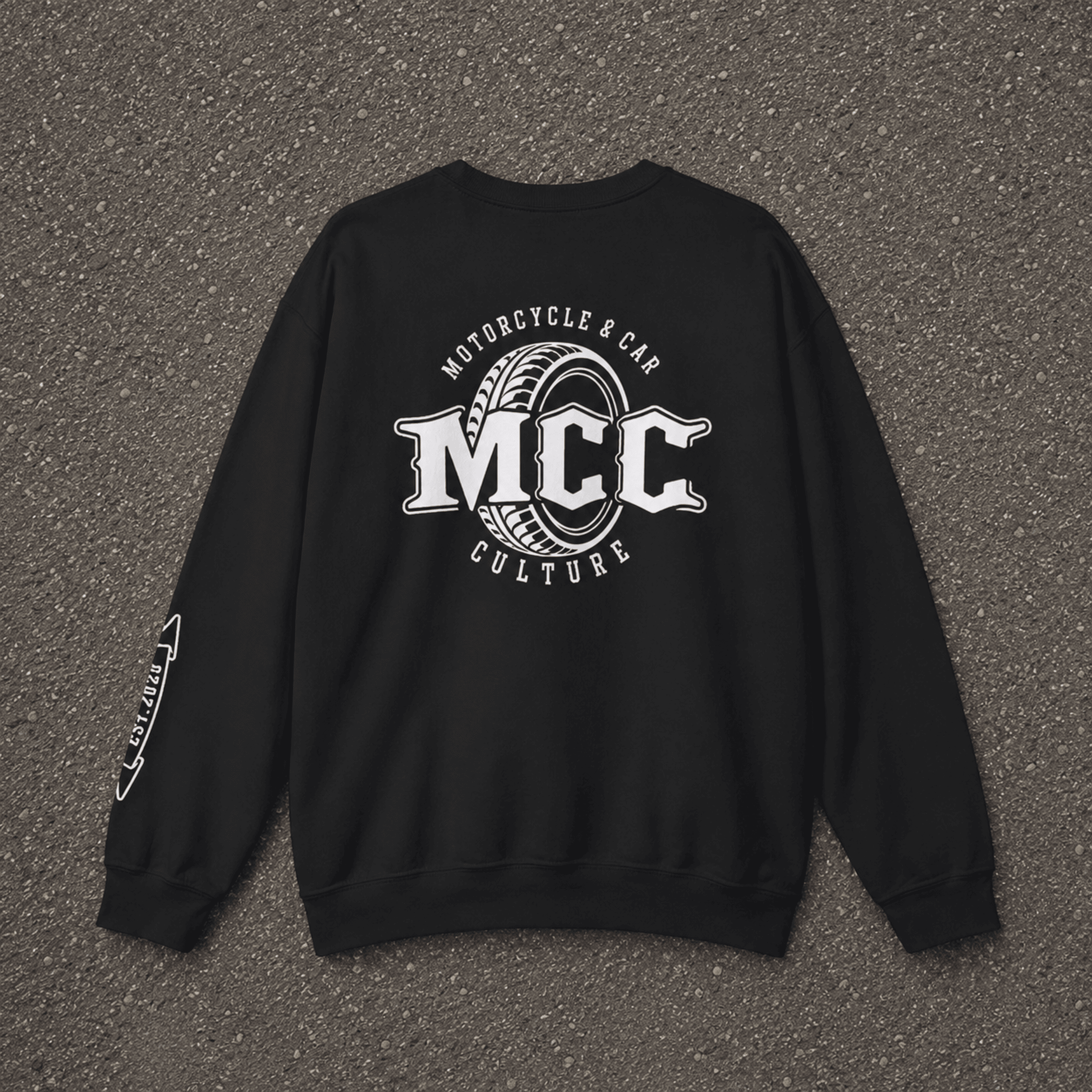 A black crewneck sweatshirt laid flat on a textured gray asphalt surface, featuring a large white graphic on the back with the text 'MCC Motorcycle & Car Culture' and a tire design, plus a smaller white graphic on the left sleeve.