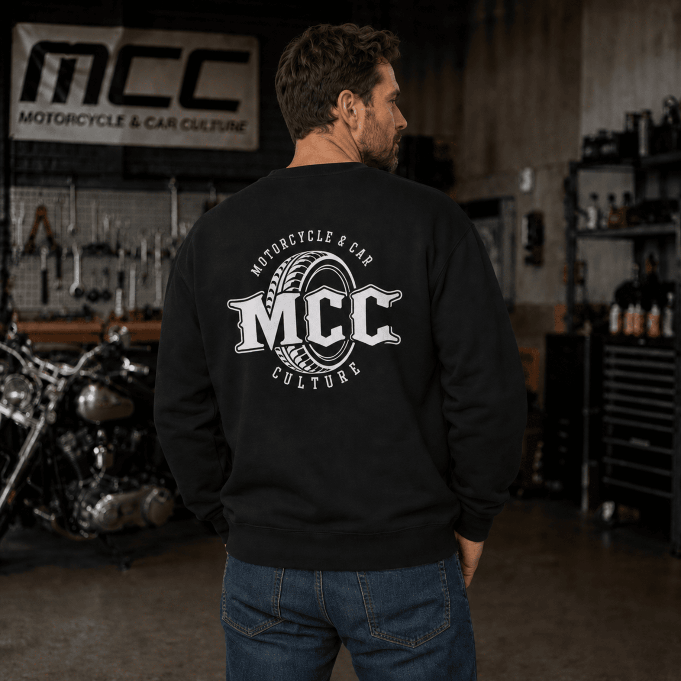 A man wearing a black crewneck sweatshirt with a large white 'MCC Motorcycle & Car Culture' graphic on the back, standing in a garage workshop environment.