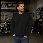 A man wearing a black crewneck sweatshirt with a white MCC Ribbon saying EST. 2026 graphic on the left sleeve, standing in a garage setting with a motorcycle and workshop equipment in the background.