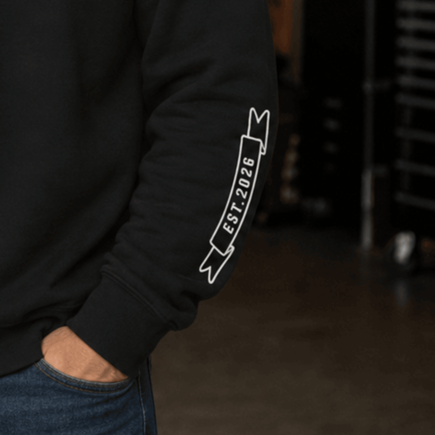 A close-up of a person wearing a black sweatshirt with a white outlined banner graphic on the left sleeve that reads 'EST. 2026'.