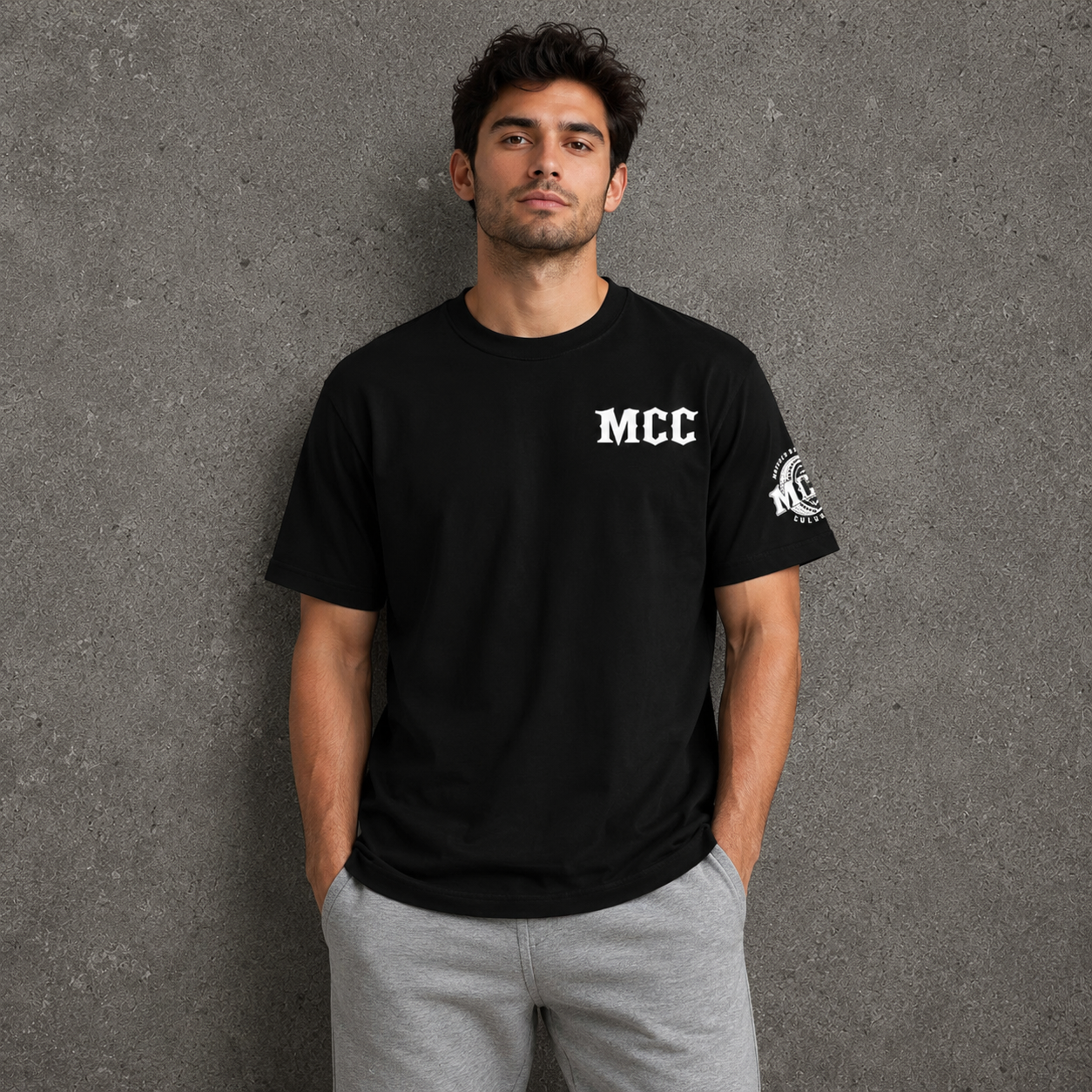 A man wearing a black short-sleeve t-shirt with 'MCC' printed in white on the left chest and a circular graphic on the left sleeve, paired with gray sweatpants, standing against a textured gray wall.
