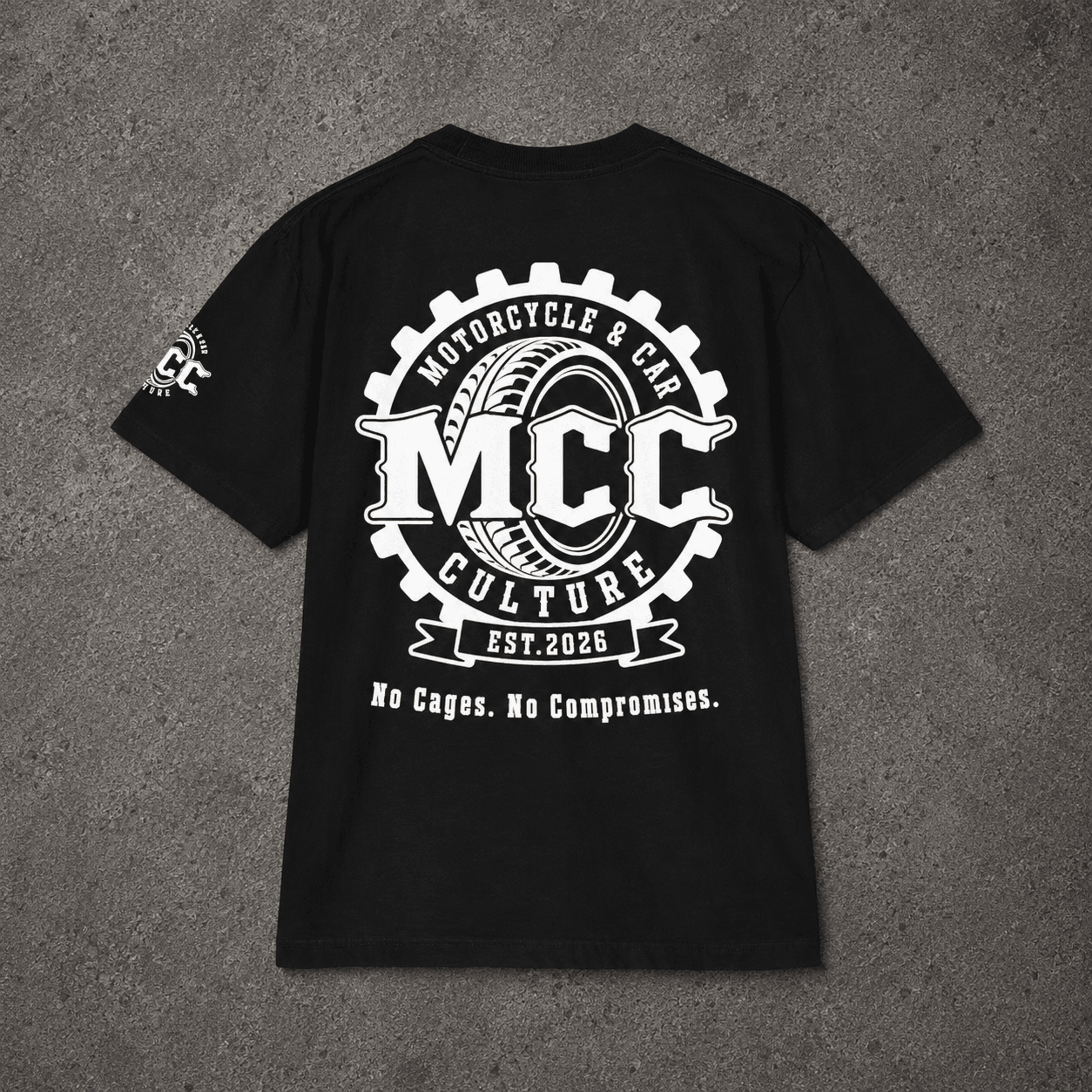 A black short-sleeve t-shirt laid flat on a textured gray background, featuring a large white graphic print on the back with the text 'MCC Motorcycle & Car Culture Est. 2026' and 'No Cages.