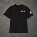A black short-sleeve t-shirt laid flat on a textured gray concrete background. The shirt features the letters 'MCC' printed in white on the left chest and a white graphic logo on the left sleeve.