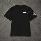 A black short-sleeve t-shirt laid flat on a textured gray concrete background. The shirt features the letters 'MCC' printed in white on the left chest and a white graphic logo on the left sleeve.