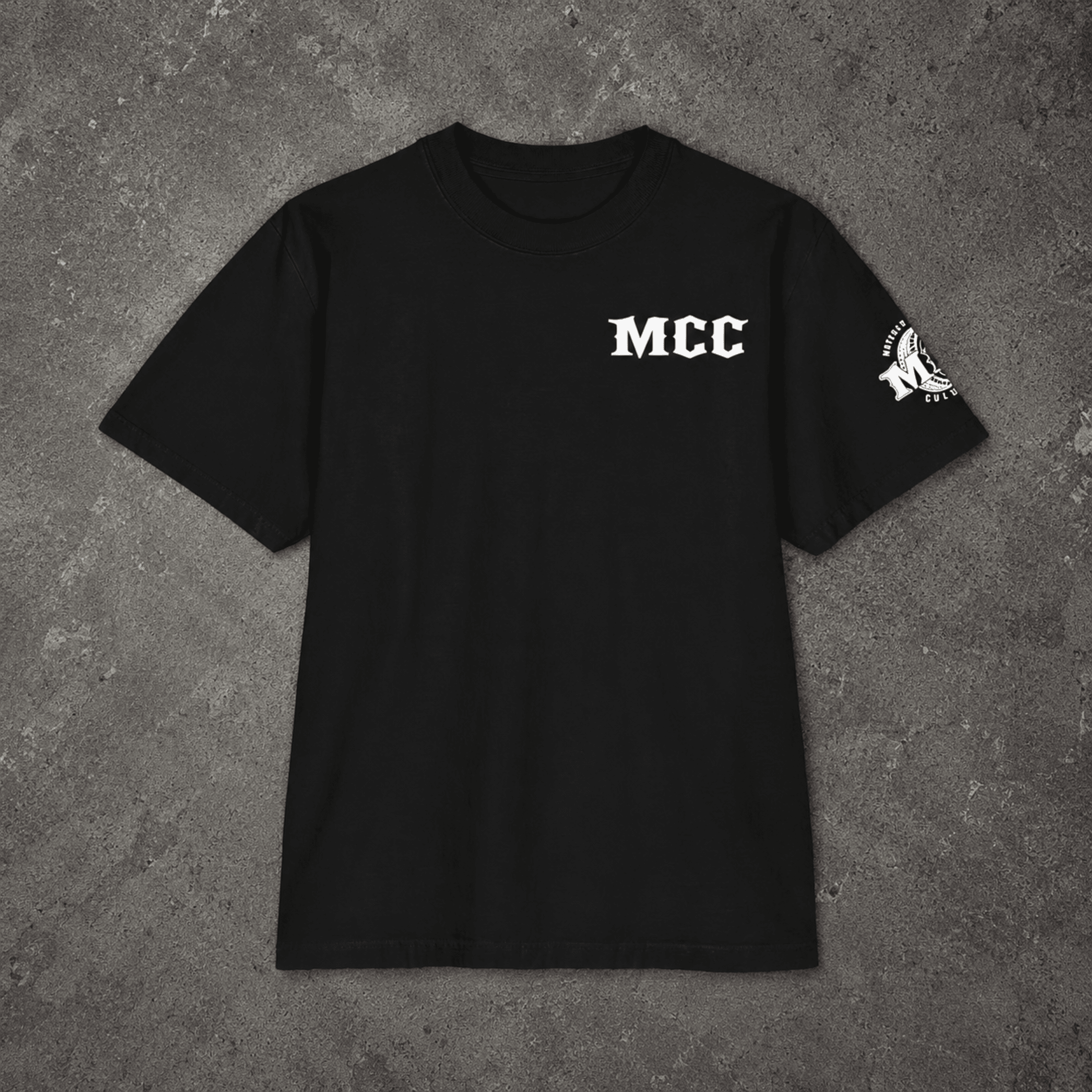 A black short-sleeve t-shirt laid flat on a textured gray concrete background. The shirt features the letters 'MCC' printed in white on the left chest and a white graphic logo on the left sleeve.