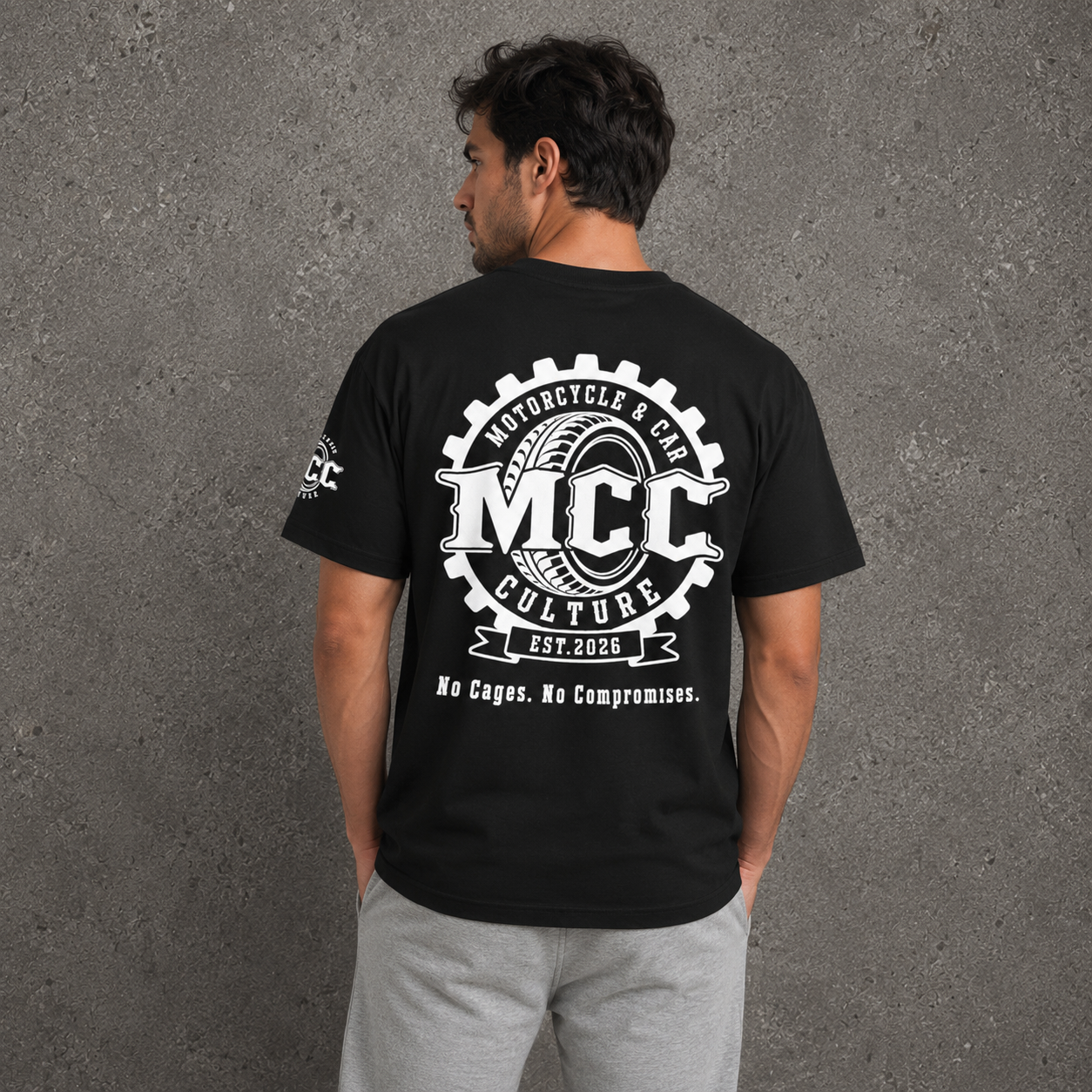 A man viewed from behind wearing a black t-shirt with a large white graphic on the back that reads 'MCC Motorcycle & Car Culture Est. 2026' and 'No Cages. No Compromises.