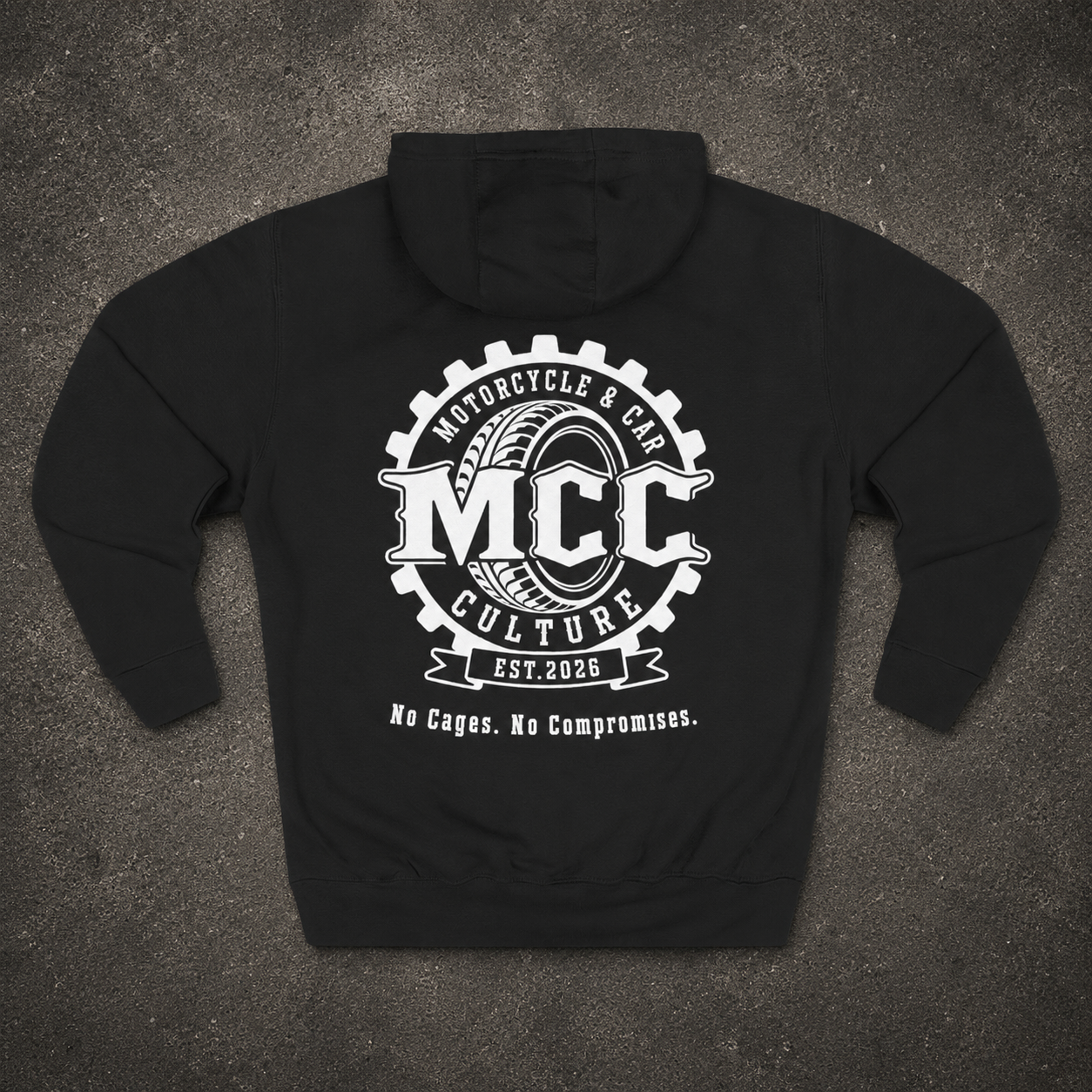 A black pullover hoodie shown from the back, laid flat on a dark textured surface. The back features a large white circular graphic with a tire and gear design, reading 'MCC Motorcycle & Car Culture' and 'No Cages. No Compromises.