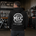 A man viewed from behind wearing a black hoodie with a large white circular 'MCC Motorcycle & Car Culture' graphic on the back, standing in a garage with a motorcycle and workshop equipment in the background.