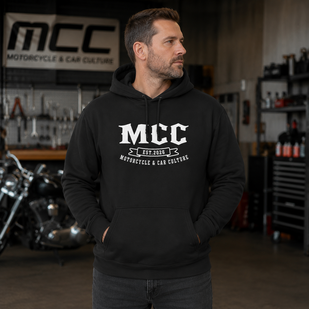 A man wearing a black pullover hoodie with a white 'MCC Motorcycle & Car Culture' graphic on the chest, standing in a garage workshop environment with a motorcycle and toolboxes in the background.