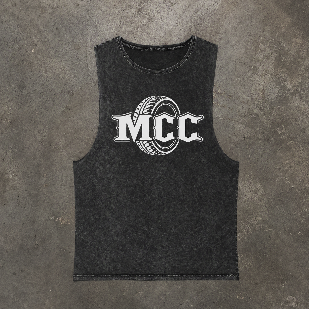 A dark gray Stonewashed sleeveless tank top featuring a white graphic print of the letters 'MCC' inside a tire design, laid flat on a textured gray concrete background.
