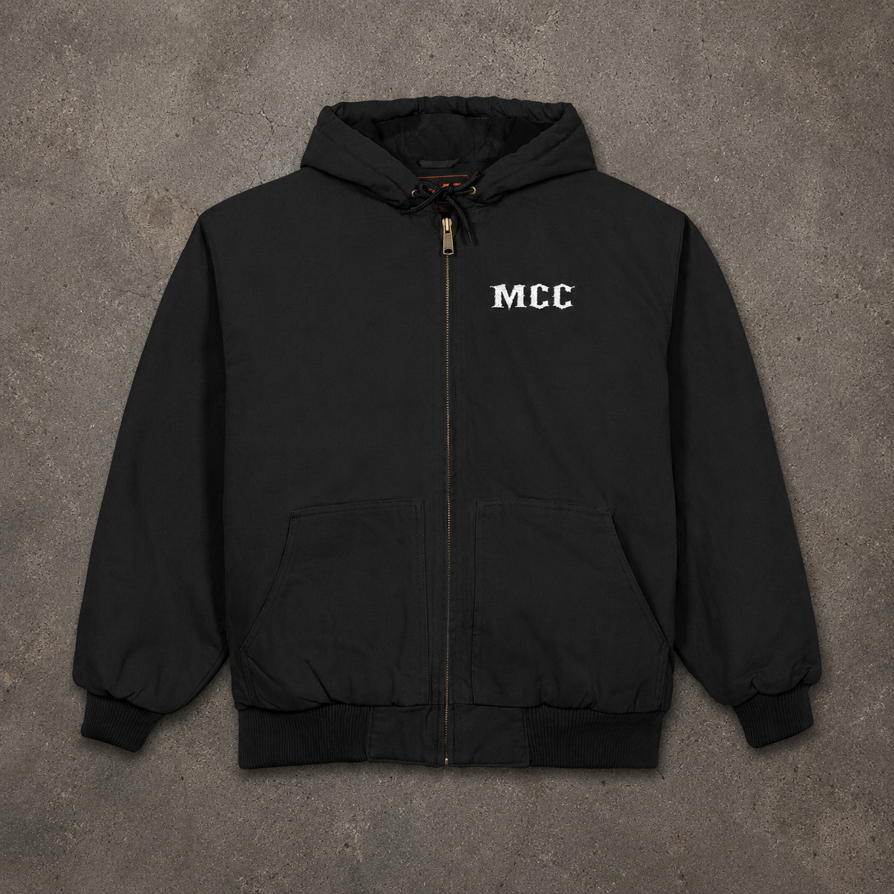 A black zip-up hooded jacket with 'MCC' printed in white on the left chest, laid flat on a textured gray concrete background.
