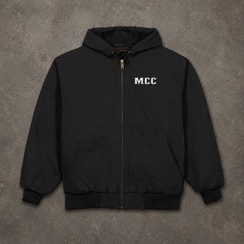 A black zip-up hooded jacket with 'MCC' printed in white on the left chest, laid flat on a textured gray concrete background.