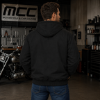 A man wearing a black hooded jacket is shown from behind in a motorcycle shop, with a motorcycle and workshop equipment visible in the background.
